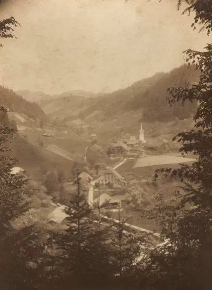 Historische Aufnahme eines idyllischen Schweizer Tals mit verstreuten Bauernhöfen, Wiesen und einem Dorf mit Kirche im Zentrum, umgeben von bewaldeten Hügeln. Die Aufnahme ist in Sepia-Tönen gehalten und von Bäumen im Vordergrund eingerahmt.