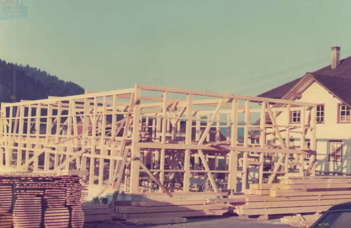 Historische Aufnahme eines Holzrahmenbaus im Entstehen, vermutlich aus den 1970er-Jahren. Im Vordergrund liegen gestapelte Holzbretter, im Hintergrund ist ein traditionelles Schweizer Wohnhaus zu sehen, umgeben von Hügeln und blauem Himmel.