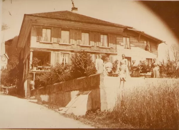 Sepiafarbenes historisches Foto eines grossen Bauernhauses mit mehreren Fenstern und Satteldach. Vor dem Haus stehen mehrere Personen, vermutlich eine Familie, umgeben von Garten und Feld. Das Bild vermittelt eine ländliche, traditionelle Atmosphäre.