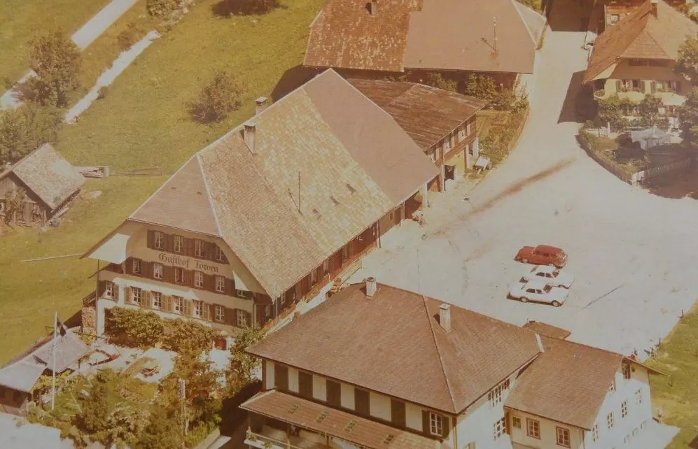 Historische Luftaufnahme des Dorfzentrums von Trub mit dem Gasthof Löwen im Vordergrund. Zu sehen sind traditionelle Emmentaler Häuser mit Schindeldächern, ein kleiner Platz mit zwei Autos und grüne Wiesen im Hintergrund.