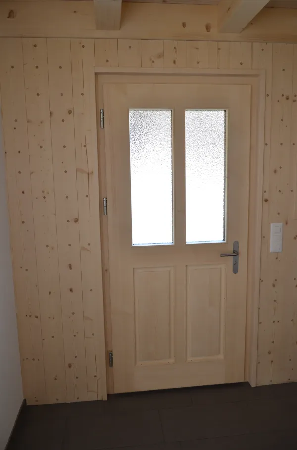 Helle Holztür mit Glaseinsätzen in einer Wandverkleidung aus naturbelassenem Holz. Die Tür hat klassische Füllungen und einen modernen Metallgriff – ein Beispiel für handwerklich gefertigte Schreinerarbeit.
