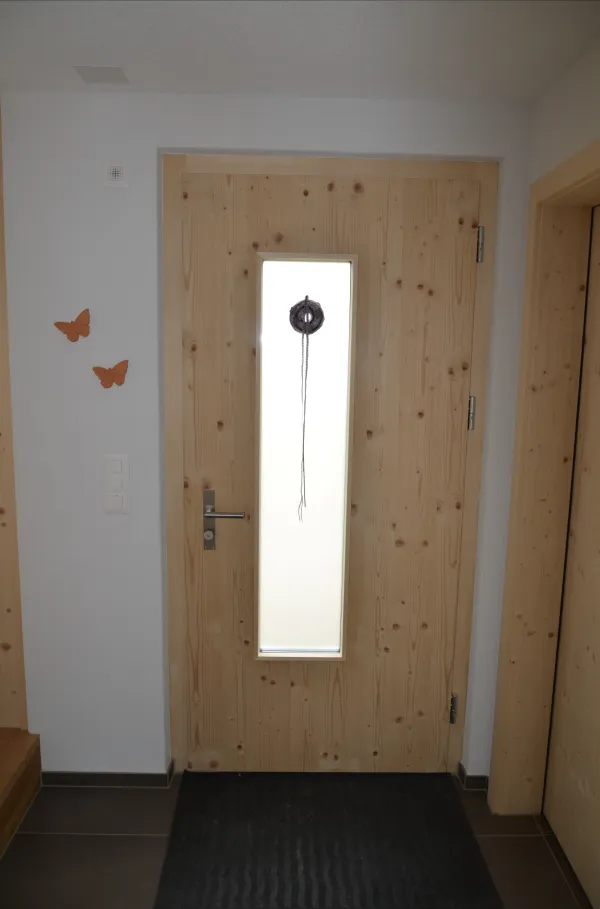 Helle Holztür mit schmalem, senkrechtem Glaseinsatz und modernem Metallgriff. Die Tür ist von einer weissen Wand umgeben, daneben sind zwei dekorative Schmetterlinge angebracht. Ein schlichtes Beispiel für zeitgemässen Innenausbau in Holz.