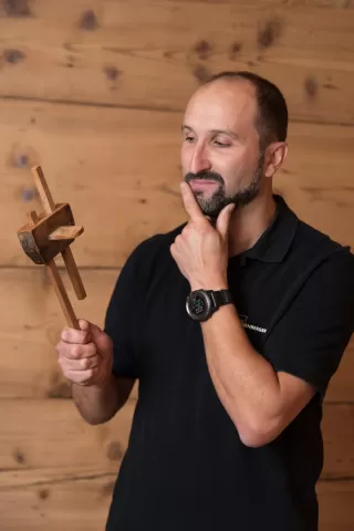 Mann in schwarzem Polohemd der Schreinerei Ellenberger betrachtet nachdenklich ein hölzernes Werkzeug vor einer Holz-Wand.