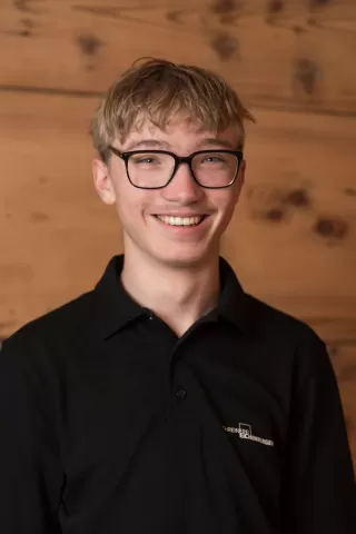 Junger Mann mit Brille und blondem Haar lächelt freundlich in die Kamera. Er trägt ein schwarzes Polohemd mit dem Logo der Schreinerei Ellenberger und steht vor einer hölzernen Wand. Das Porträt vermittelt einen sympathischen, offenen Eindruck und spiegelt die Freude an der handwerklichen Arbeit wider.“